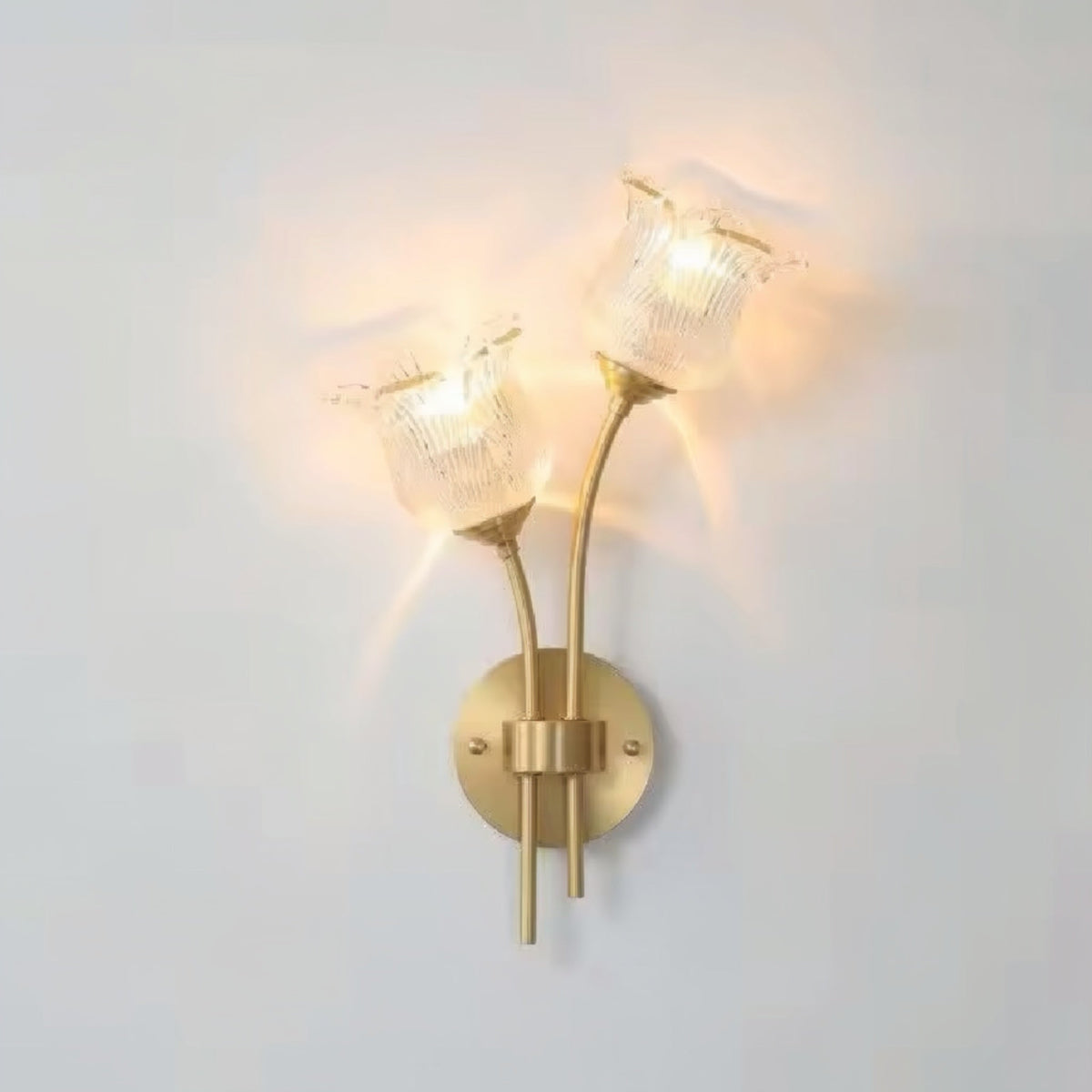 Flora Luxe botanical brass double-head wall sconce with clear ribbed floral glass shades illuminated against a white wall, angled left — LivDecor