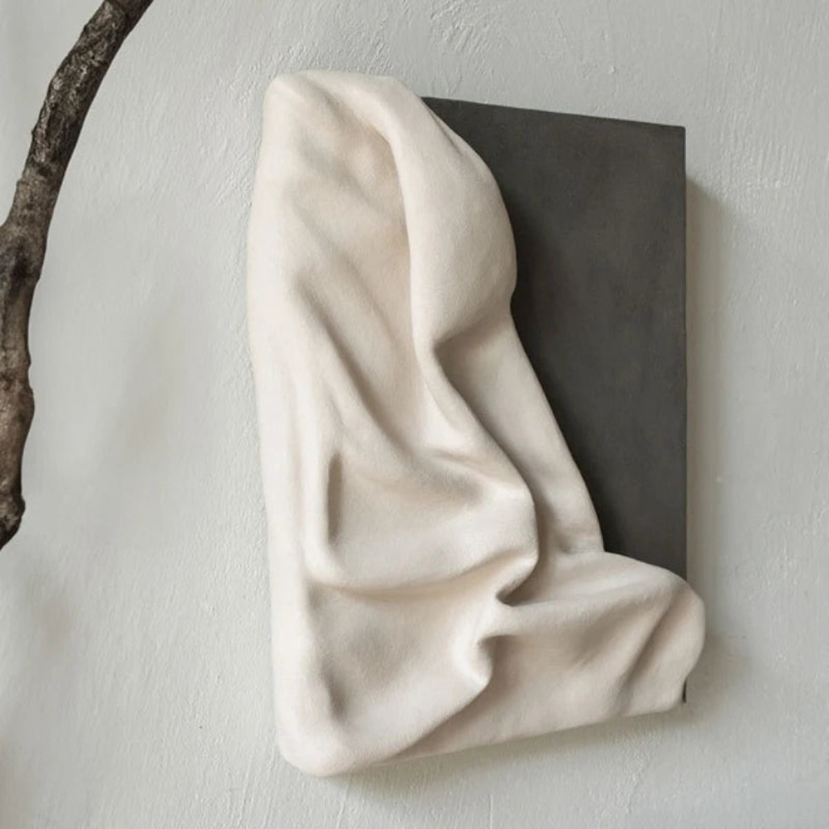3D draped cream fabric resin wall sculpture on a charcoal grey panel photographed at an angle with a sculptural bare branch in the foreground — LivDecor