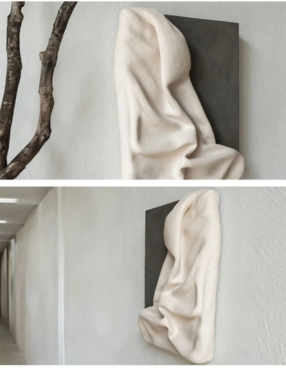 Two lifestyle shots of 3D draped cream resin wall sculpture on charcoal panel — top styled with a sculptural bare branch, bottom displayed in a bright minimalist hallway — LivDecor