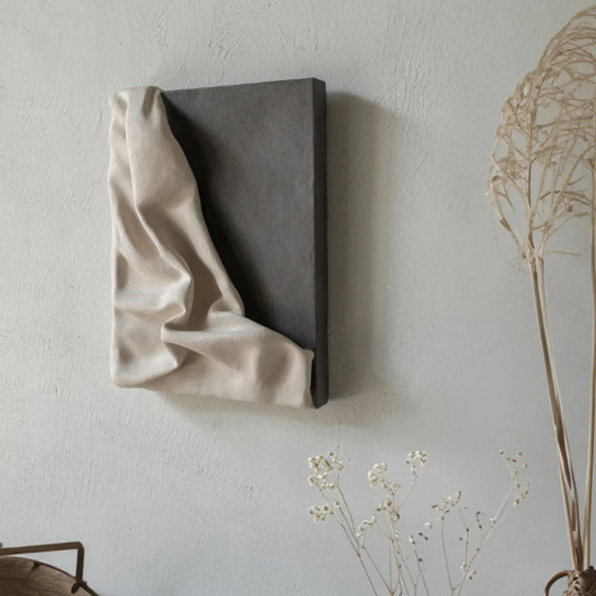 3D draped cream fabric resin wall sculpture on a charcoal grey panel mounted on a textured white wall, styled with dried botanical stems in the foreground — LivDecor