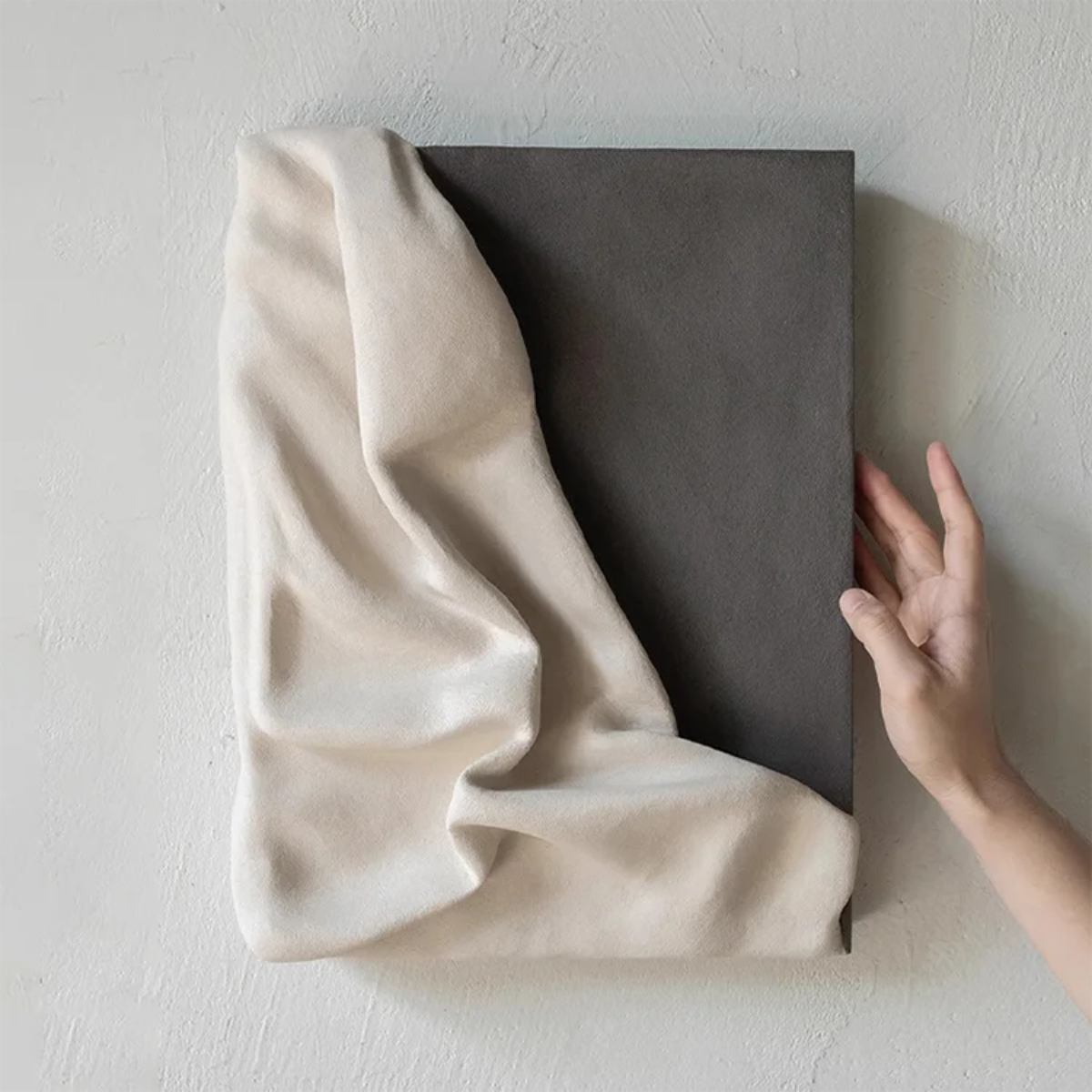 3D draped cream fabric resin wall sculpture on a charcoal grey panel with a hand placed beside it to show scale — LivDecor