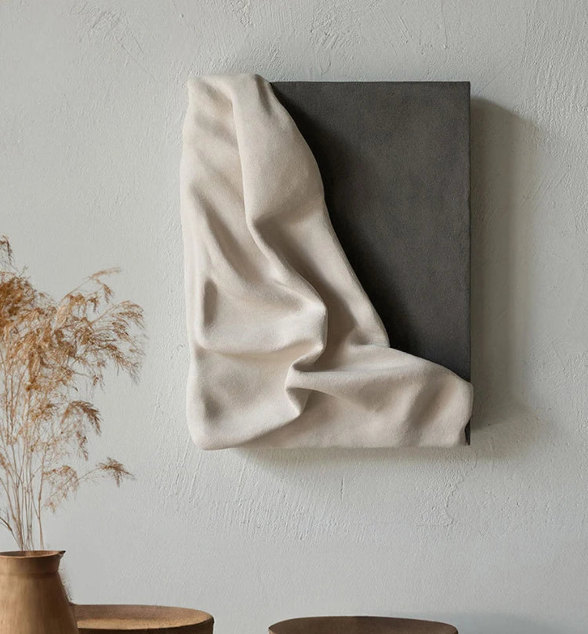 3D draped cream fabric resin wall sculpture on a charcoal grey panel mounted on a textured white wall, styled with dried pampas grass in a terracotta vase and wooden side table — LivDecor
