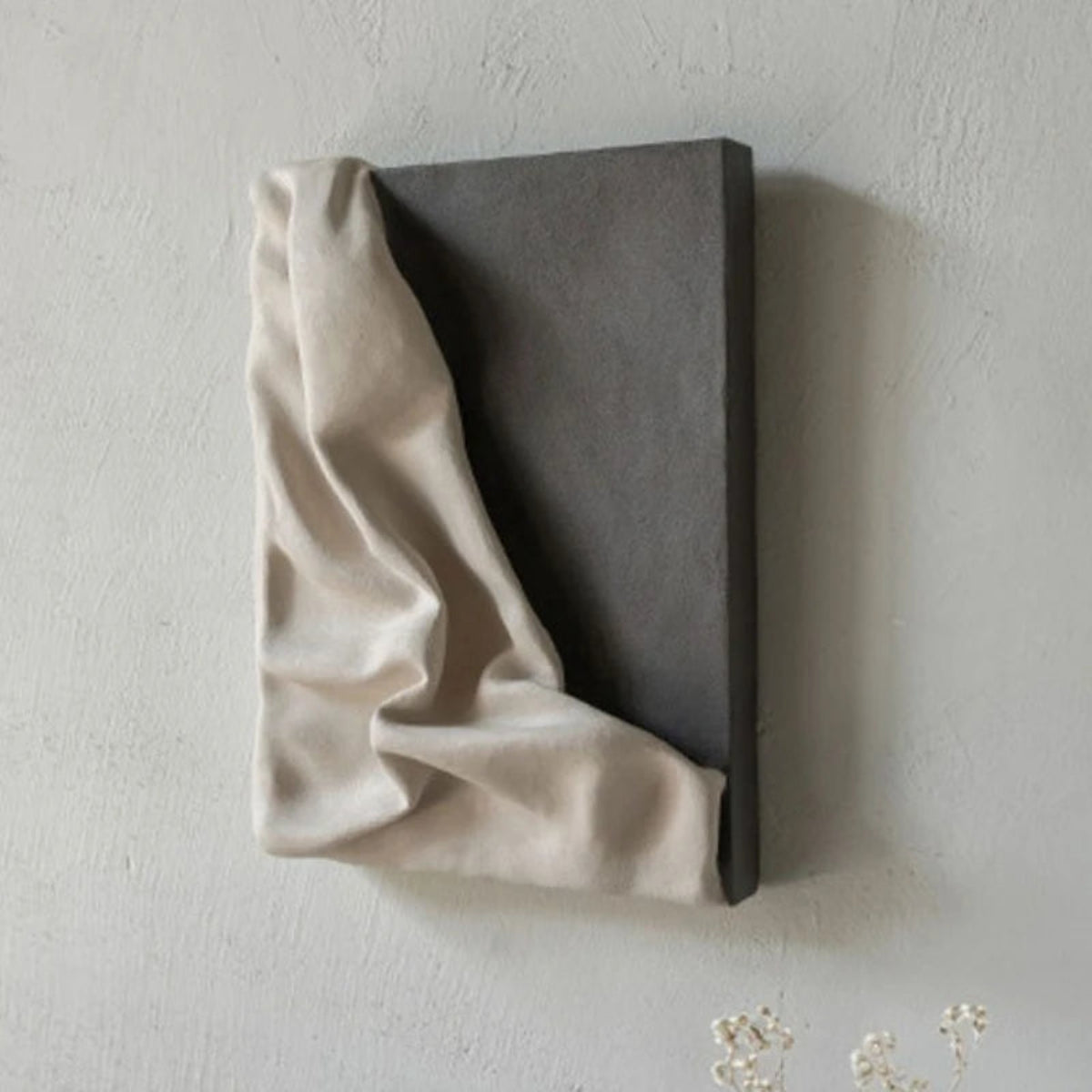 3D draped cream fabric resin wall sculpture on a charcoal grey panel photographed at a side angle showing depth and shadow, with small white floral sprigs at the base — LivDecor