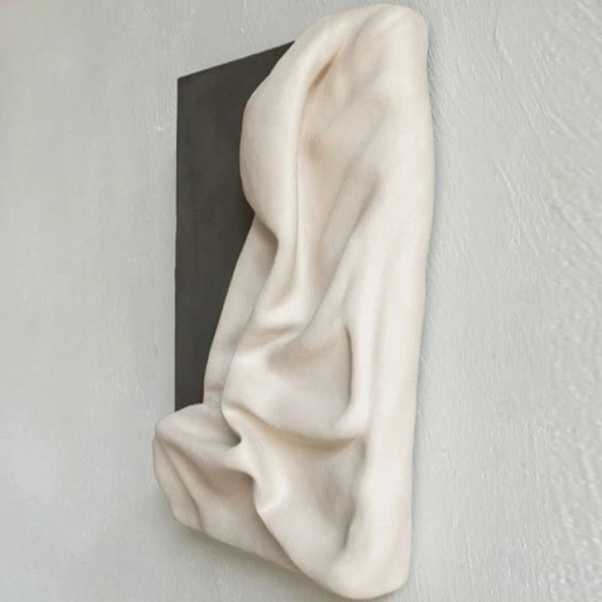 Side profile view of 3D draped cream fabric resin wall sculpture on a charcoal grey panel showing the full sculptural depth and projection from the wall — LivDecor