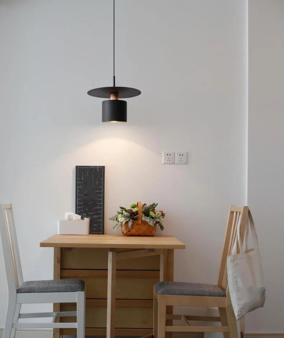 Aero Slim cylinder pendant light in matte black with copper accent lit over a small pine breakfast table with mismatched chairs and wicker flower basket against a white wall — LivDecor