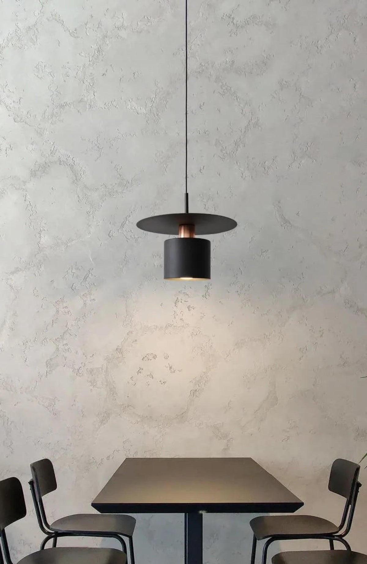 Aero Slim cylinder pendant light in matte black with copper accent lit over a square black dining table with black school chairs against a raw plaster wall casting warm downward glow — LivDecor

