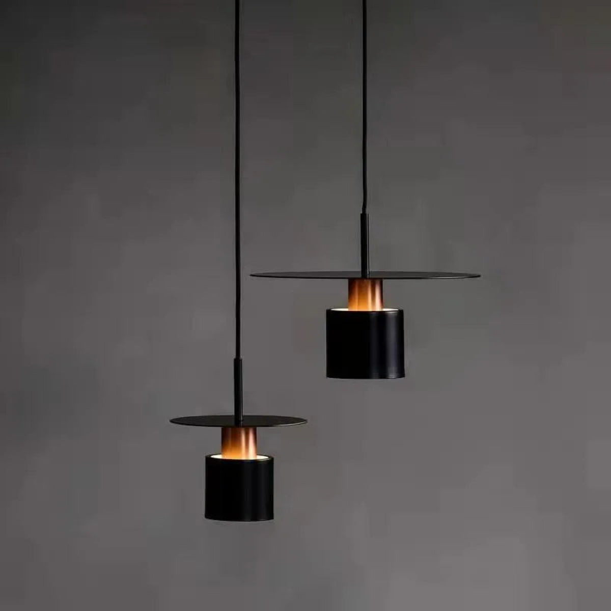 Two Aero Slim cylinder pendant lights in matte black with glowing copper accent collar hung at staggered heights and illuminated against a dark charcoal grey background — LivDecor
