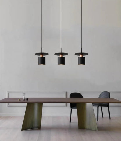 Three Aero Slim cylinder pendant lights in matte black with copper accent hung in a linear row over a walnut and olive green trestle dining table with black shell chairs against a white panelled wall — LivDecor
