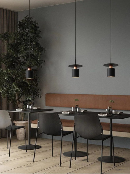 Three Aero Slim cylinder pendant lights in matte black with copper accent hung at staggered heights over bistro tables with cork banquette seating against slate blue walls in a moody restaurant setting — LivDecor
