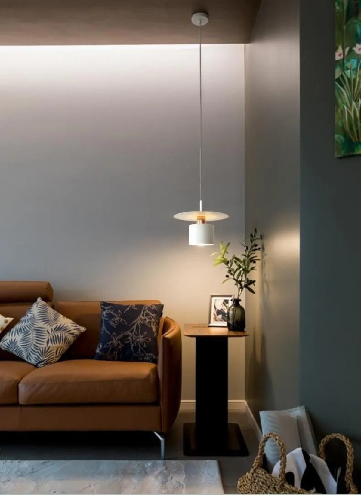 Aero Slim cylinder pendant light in matte white with copper accent lit over a pedestal side table with vase and framed art beside a tan leather sofa against moody teal-grey walls — LivDecor
