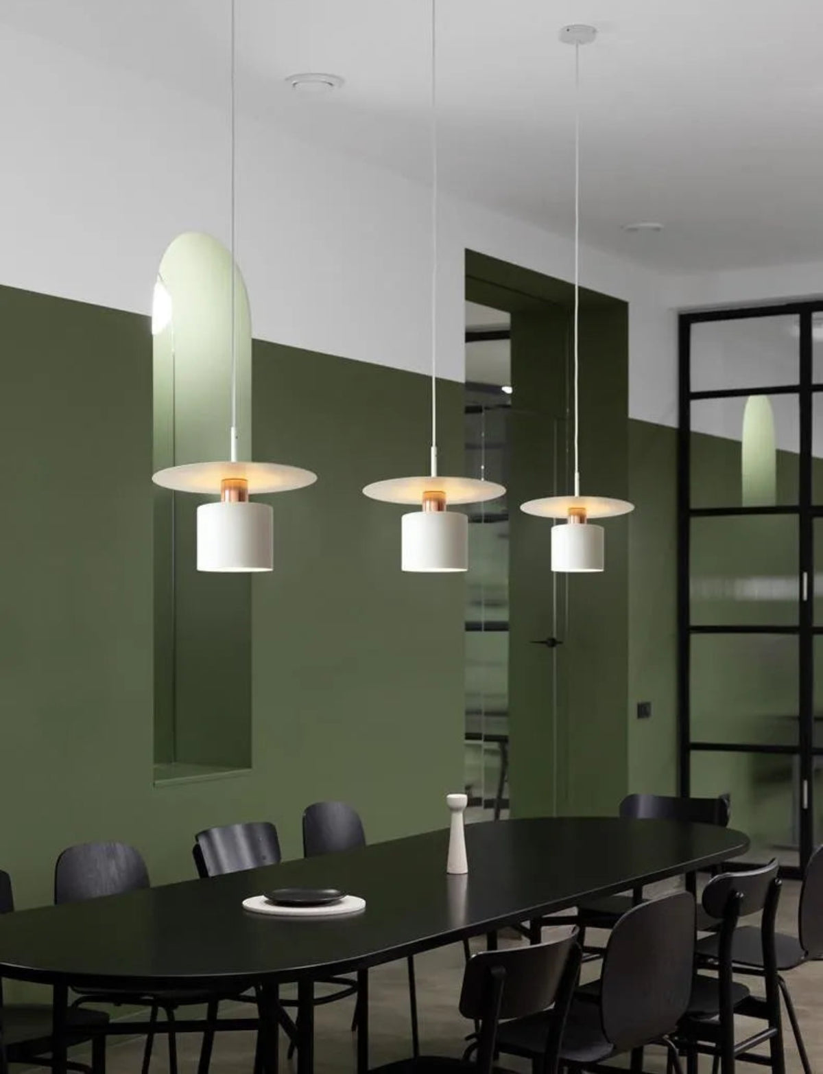 Three Aero Slim cylinder pendant lights in matte white with copper accent hung in a linear row over a large oval black dining table against deep forest green walls with black steel frame doors — LivDecor
