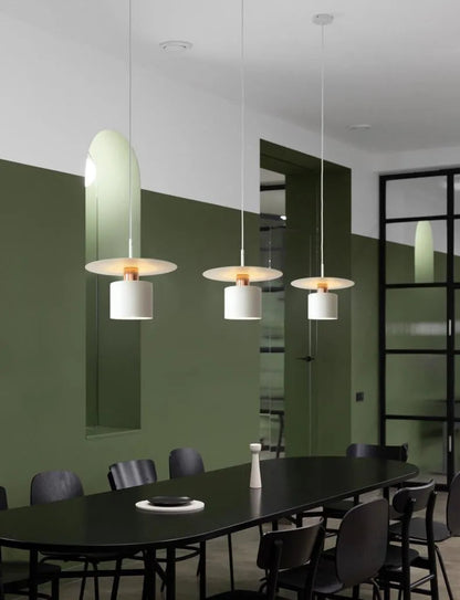 Three Aero Slim cylinder pendant lights in matte white with copper accent hung in a linear row over a large oval black dining table against deep forest green walls with black steel frame doors — LivDecor
