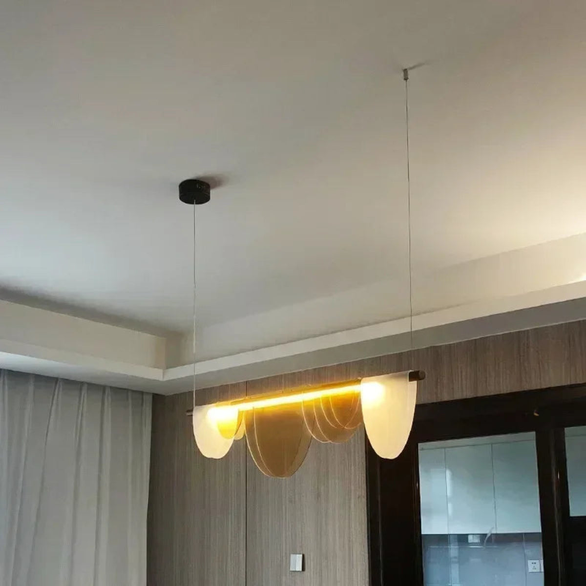 Amber acrylic petal pendant light installed in a real room showing the black ceiling canopy, steel cable drop, and warm LED glow against a neutral ceiling — LivDecor