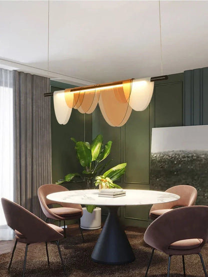 Amber acrylic petal pendant light illuminated above a round marble dining table with blush velvet chairs against deep forest green panelled walls with a tropical plant and sheer curtains — LivDecor
