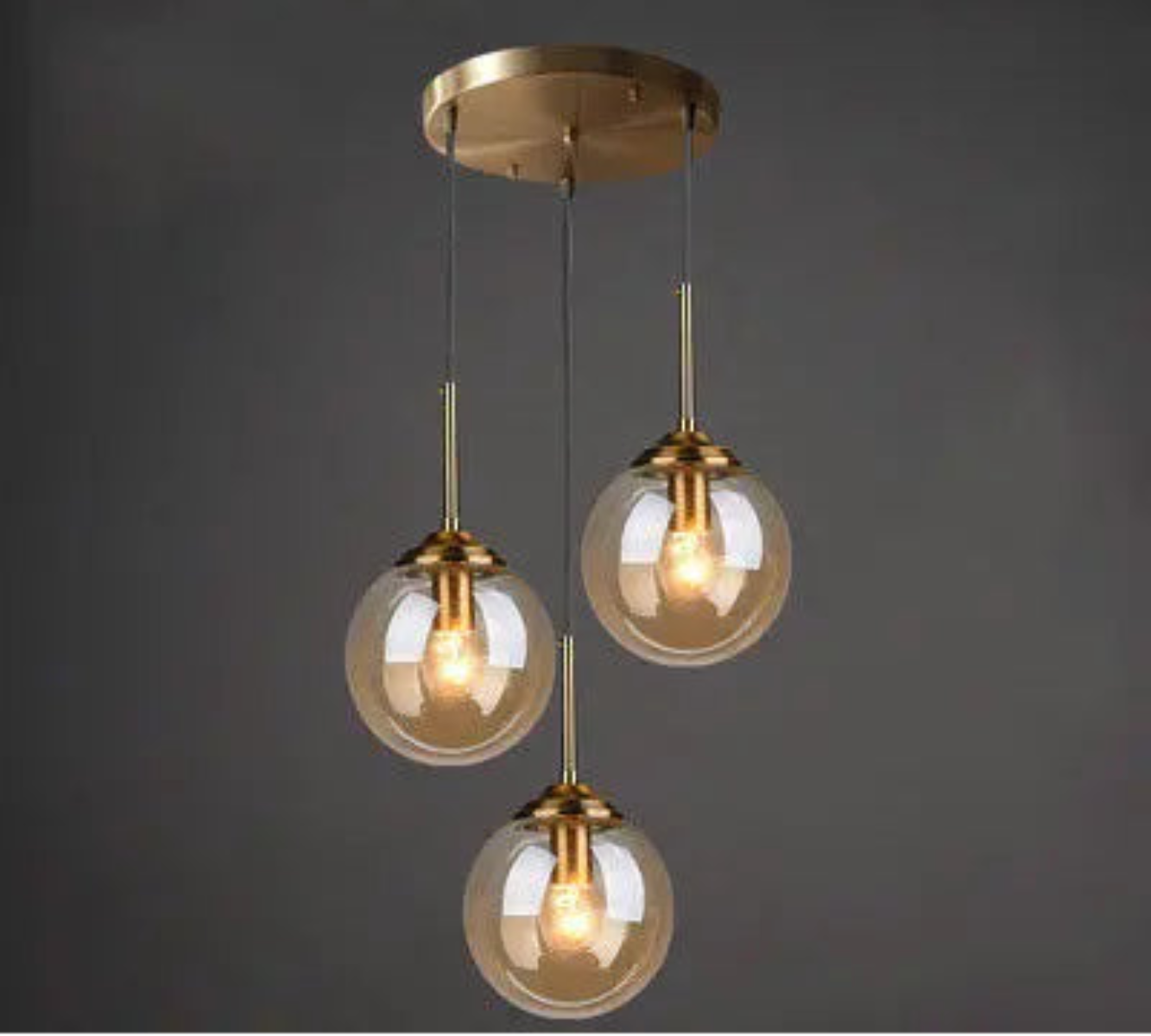 Three amber glass globe pendant lights on staggered gold cords and round ceiling mount in a cluster configuration against a dark grey background — LivDecor
