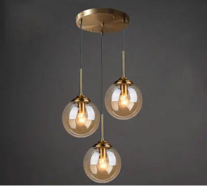 Three amber glass globe pendant lights on staggered gold cords and round ceiling mount in a cluster configuration against a dark grey background — LivDecor

