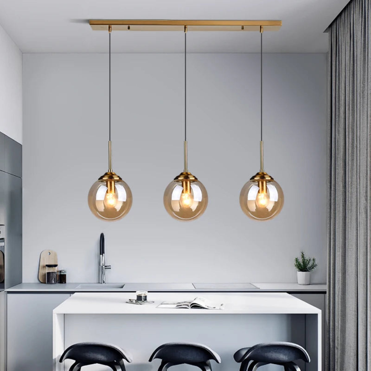 Three amber glass globe pendant lights on a gold linear bar mount hanging above a white kitchen island with black bar stools in a modern grey kitchen — LivDecor