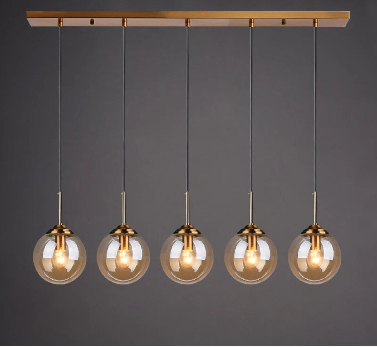 Five amber glass globe pendant lights evenly spaced on gold cords and rectangular bar ceiling mount in a linear row against a dark grey background — LivDecor
