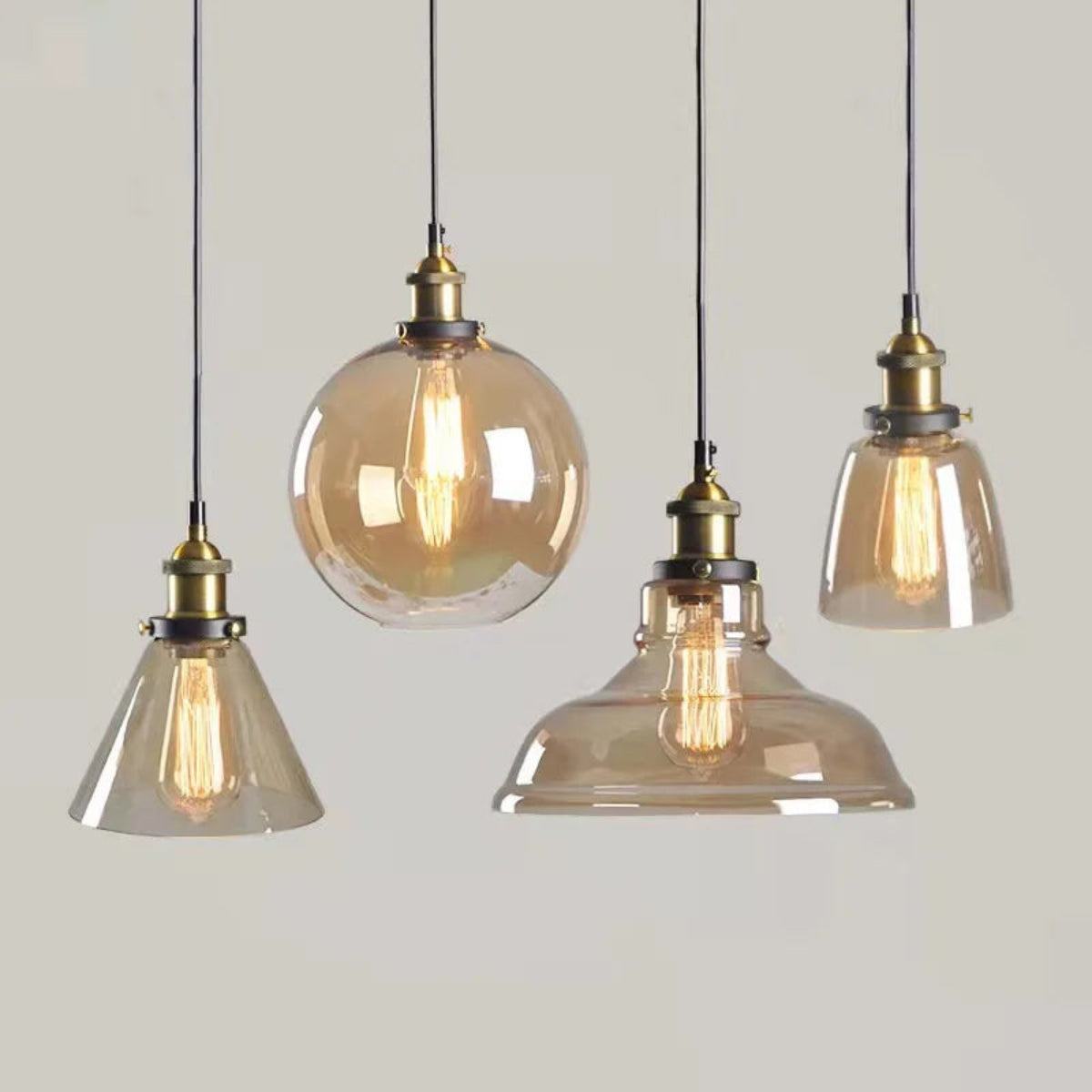 Four amber glass pendant lights in cone, globe, dome and bell shapes with brass fittings and glowing Edison filaments displayed against a neutral grey background — LivDecor