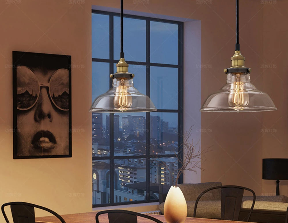 Two clear glass barn pendant lights with antique brass sockets and Edison filament bulbs hanging in a moody urban loft dining room with city skyline view at dusk — LivDecor
