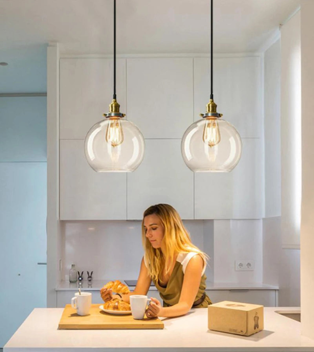 Two clear glass globe pendant lights with antique brass sockets and Edison filament bulbs above a woman enjoying breakfast at a modern white kitchen island — LivDecor
