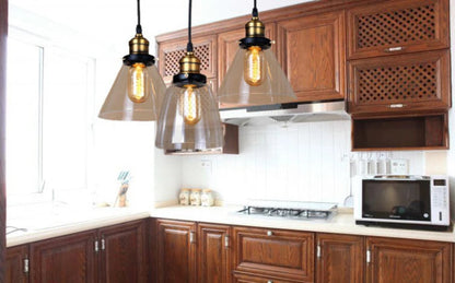 Three clear glass cone pendant lights with antique brass and black sockets and Edison filament bulbs hanging above a warm walnut wood kitchen with white countertops — LivDecor
