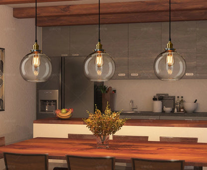 Three clear glass globe pendant lights with antique brass sockets and Edison filament bulbs hanging over a rustic wood dining table in a modern kitchen — LivDecor
