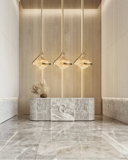 Three amber wave glass geometric pendant lights on gold stems hanging above a marble reception desk with pampas grass decor in a luxury hotel lobby with warm wood paneling and marble floors — LivDecor