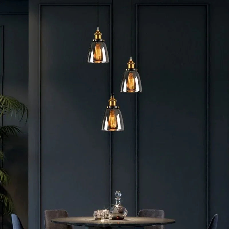 Three smoky gray glass bell pendant lights with antique brass sockets and Edison filament bulbs cascading at varying heights above a round dining table against a dark teal paneled wall — LivDecor