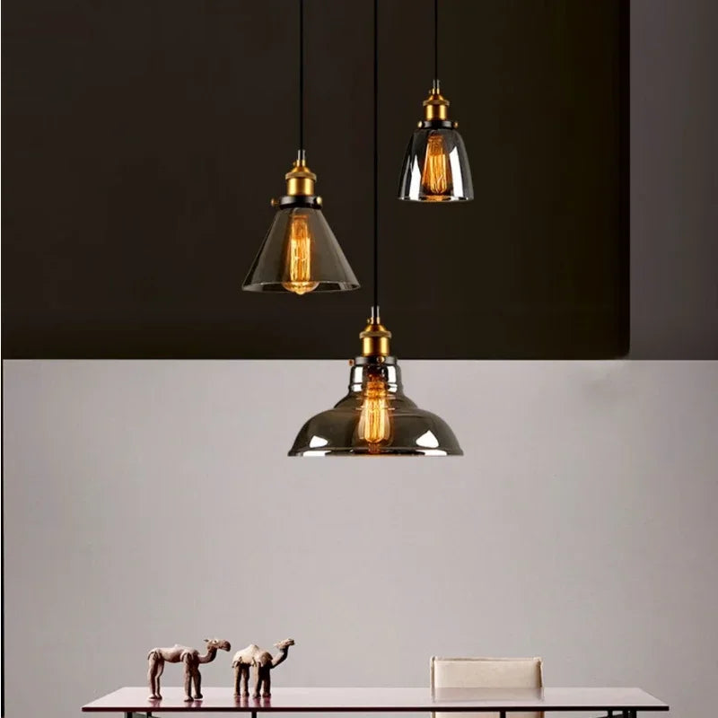Three smoky gray glass cone and bell pendant lights with antique brass sockets and Edison filament bulbs hanging above a minimalist dining table against a split dark and light background — LivDecor