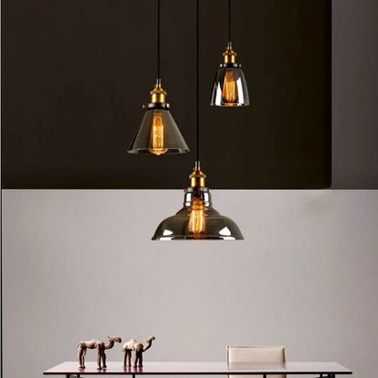 Three smoky gray glass cone and bell pendant lights with antique brass sockets and Edison filament bulbs hanging above a minimalist dining table against a split dark and light background — LivDecor