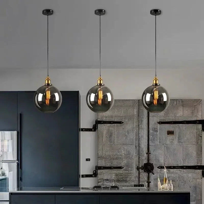 Three smoky gray glass globe pendant lights with antique brass sockets and Edison filament bulbs hanging over a dark industrial kitchen island with navy cabinetry and stone tile wall — LivDecor