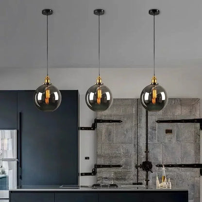 Three smoky gray glass globe pendant lights with antique brass sockets and Edison filament bulbs hanging over a dark industrial kitchen island with navy cabinetry and stone tile wall — LivDecor