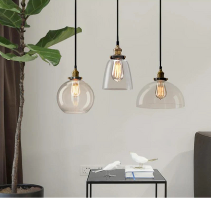Three clear glass pendant lights in globe, cone, and dome styles with antique brass sockets and Edison filament bulbs above a minimalist living room corner with a fiddle leaf fig plant — LivDecor
