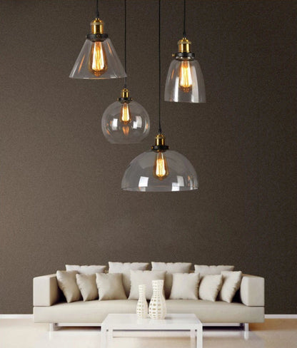 Four clear glass pendant lights in mixed styles — cone, bell, globe, and dome — with antique brass sockets and Edison filament bulbs cascading at varying heights above a modern neutral living room sofa — LivDecor