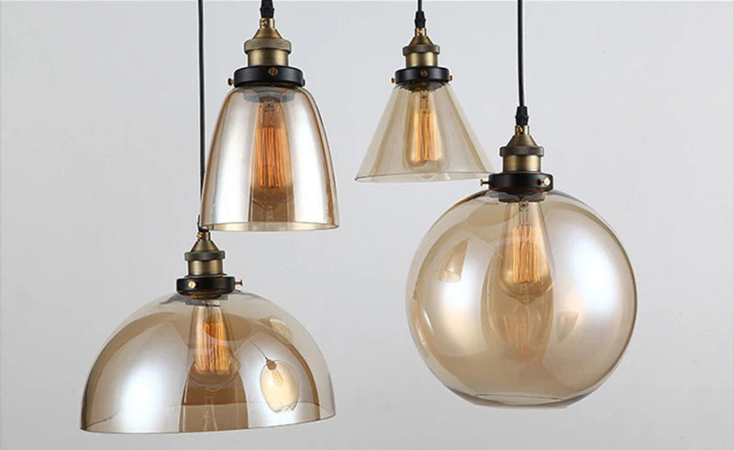 Close-up detail of four amber iridescent glass pendant lights — bell, cone, dome, and globe — with antique brass sockets and glowing Edison filament bulbs on black cords — LivDecor
