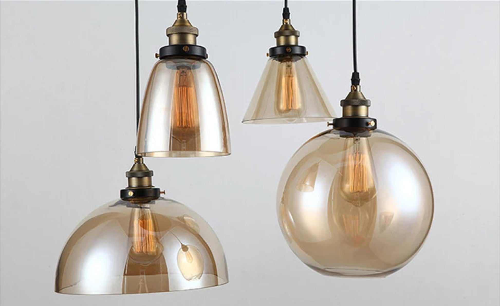 Close-up detail of four amber iridescent glass pendant lights — bell, cone, dome, and globe — with antique brass sockets and glowing Edison filament bulbs on black cords — LivDecor
