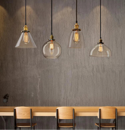 Four clear glass pendant lights in cone, globe, bell, and dome styles with antique brass sockets and Edison filament bulbs hanging in a row above a wooden bar counter against a gray wood-grain wall — LivDecor
