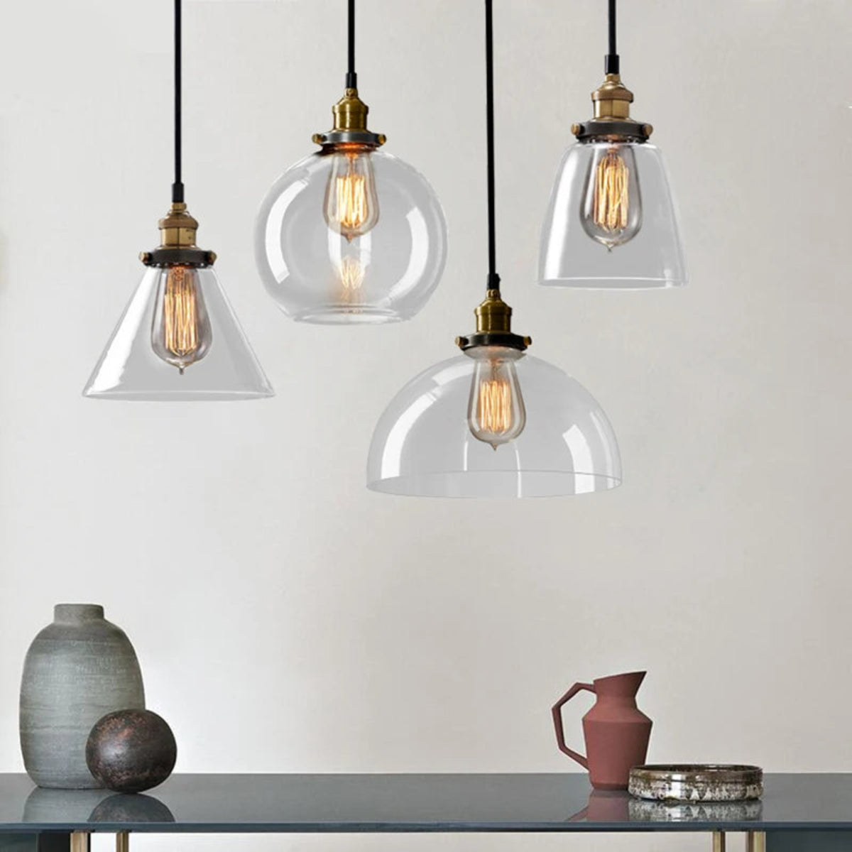 Four clear glass pendant lights — cone, globe, bell, and dome styles — with antique brass sockets and Edison filament bulbs above a styled shelf with ceramic vases and a terracotta jug against a warm white wall — LivDecor
