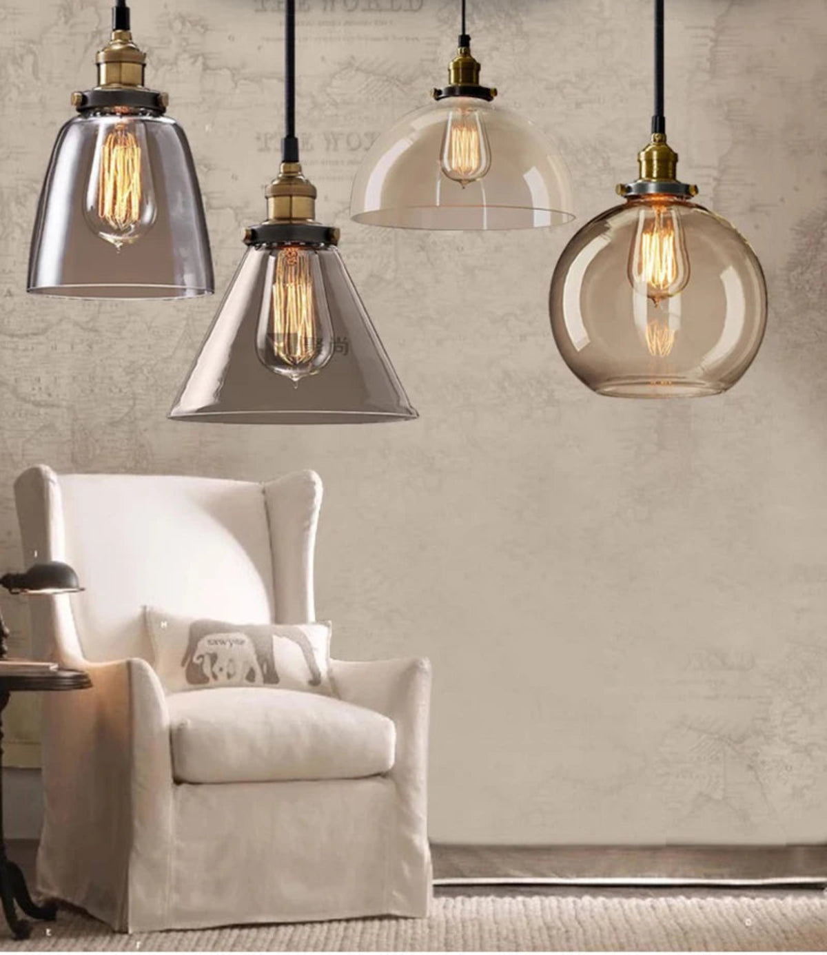 Four mixed clear and smoky glass pendant lights — bell, cone, dome, and globe — with antique brass sockets and Edison filament bulbs above a white wingback armchair against a rustic plaster wall — LivDecor
