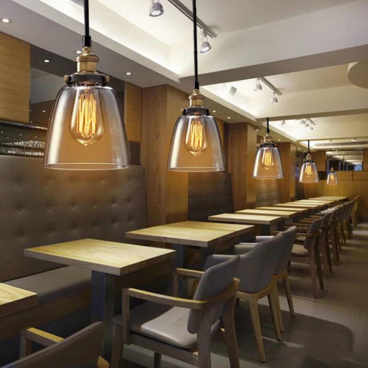 Row of clear glass bell pendant lights with antique brass sockets and Edison filament bulbs hanging above wooden dining tables in a warm modern restaurant or café interior — LivDecor
