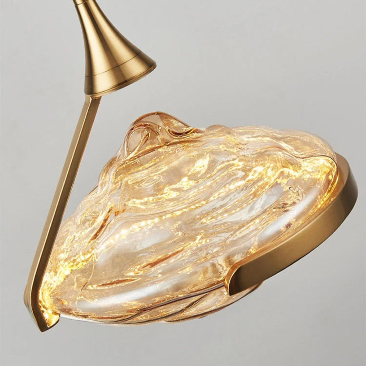 Close-up detail of an amber wave glass geometric pendant light glowing from within with swirling glass texture and brushed gold stem and ring hardware — LivDecor
