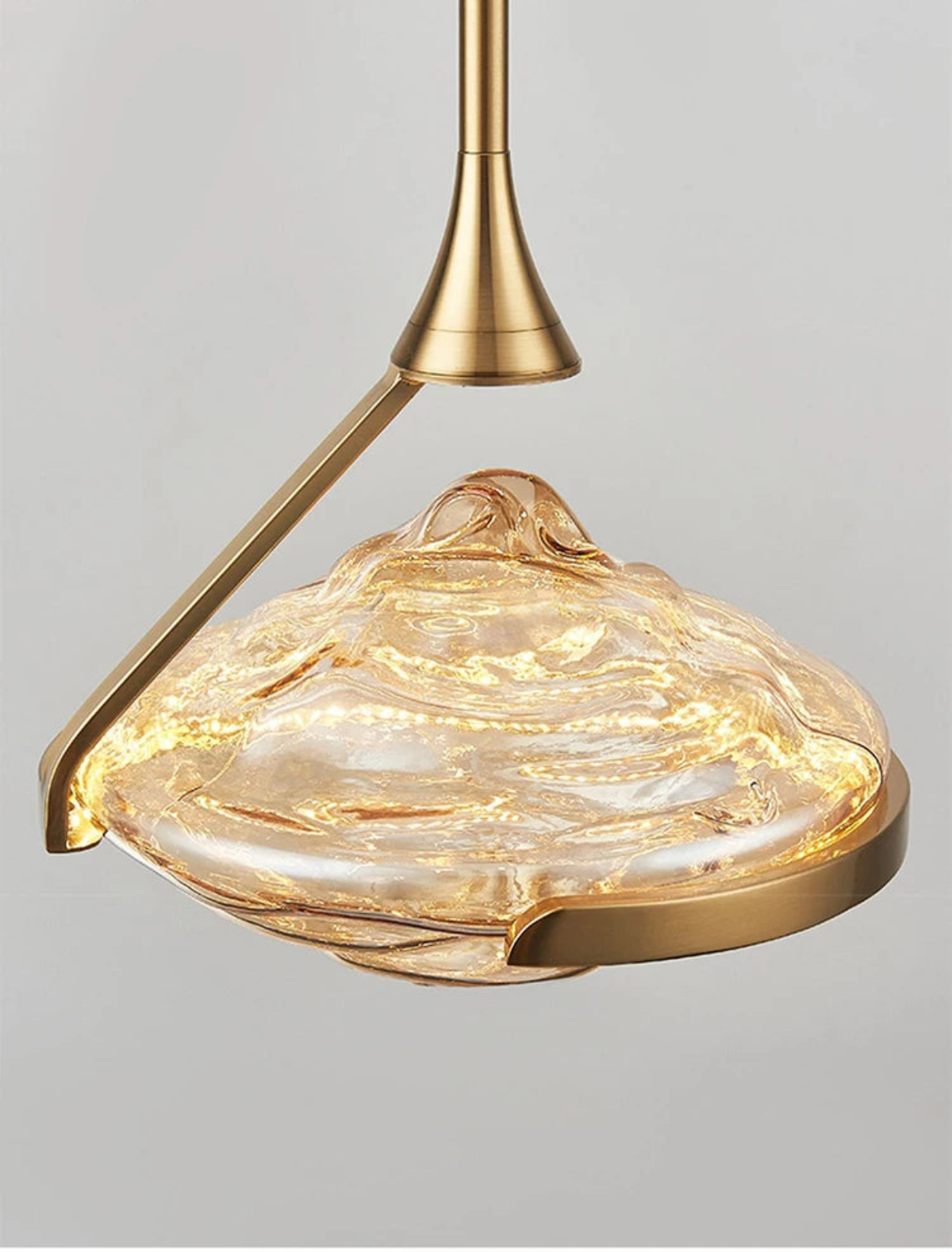 Extreme close-up of an amber wave glass geometric pendant light glowing warmly from within showing swirling glass texture and brushed gold arm and ring hardware against a grey background — LivDecor