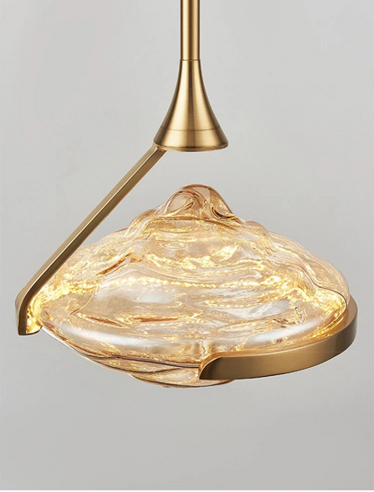 Extreme close-up of an amber wave glass geometric pendant light glowing warmly from within showing swirling glass texture and brushed gold arm and ring hardware against a grey background — LivDecor