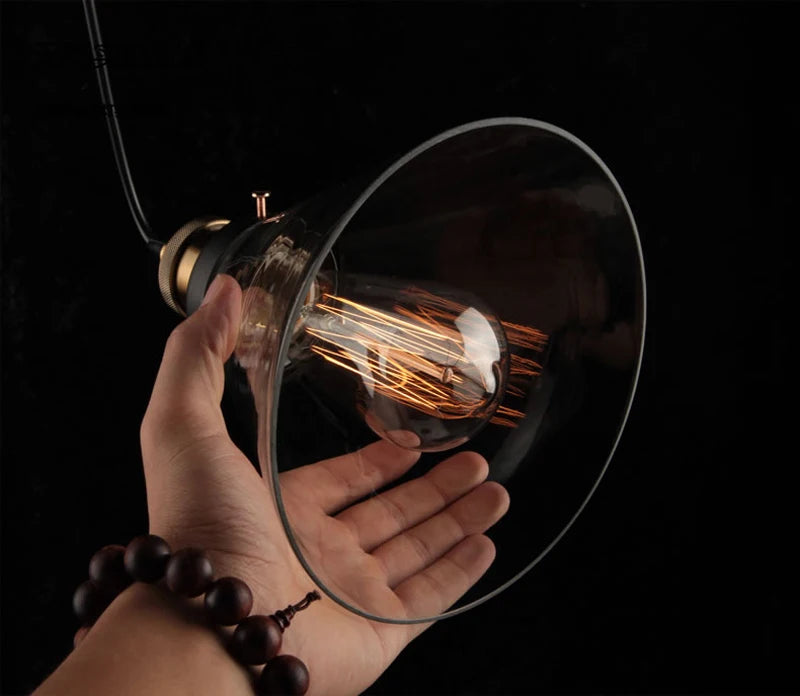 Hand holding a single clear wide flare glass pendant light with antique brass socket and glowing Edison filament bulb on black cord against a dark background — LivDecor
