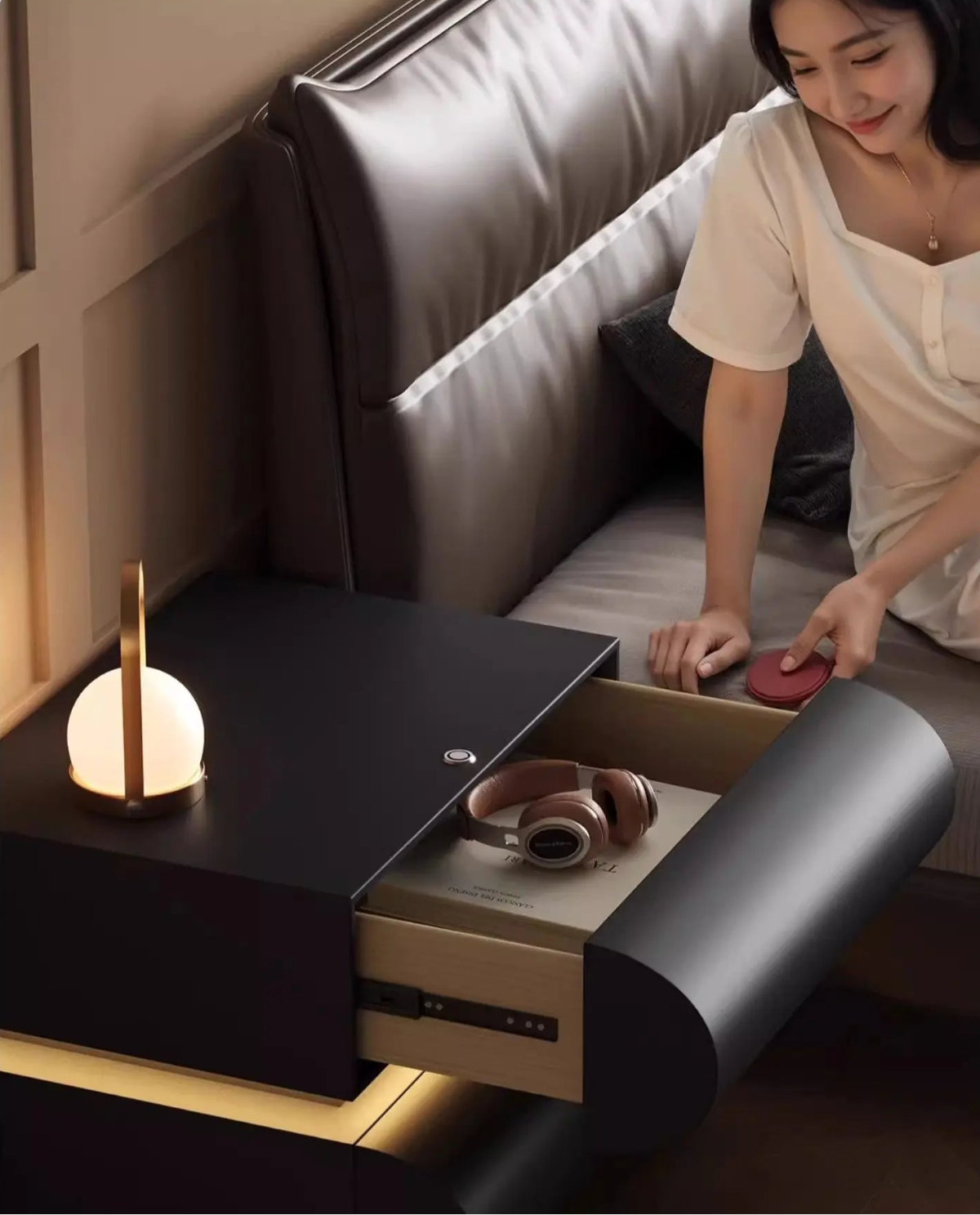 Aura Smart LED luxury nightstand in black with drawer open showing headphones and book storage, model reaching from bed in a warm moody bedroom with LED base glow — LivDecor
