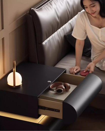 Aura Smart LED luxury nightstand in black with drawer open showing headphones and book storage, model reaching from bed in a warm moody bedroom with LED base glow — LivDecor
