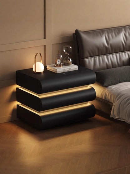 Aura Smart LED luxury nightstand in black with dual warm LED strip glow styled with a portable lamp and hourglass beside a dark leather bed in a moody luxury bedroom — LivDecor
