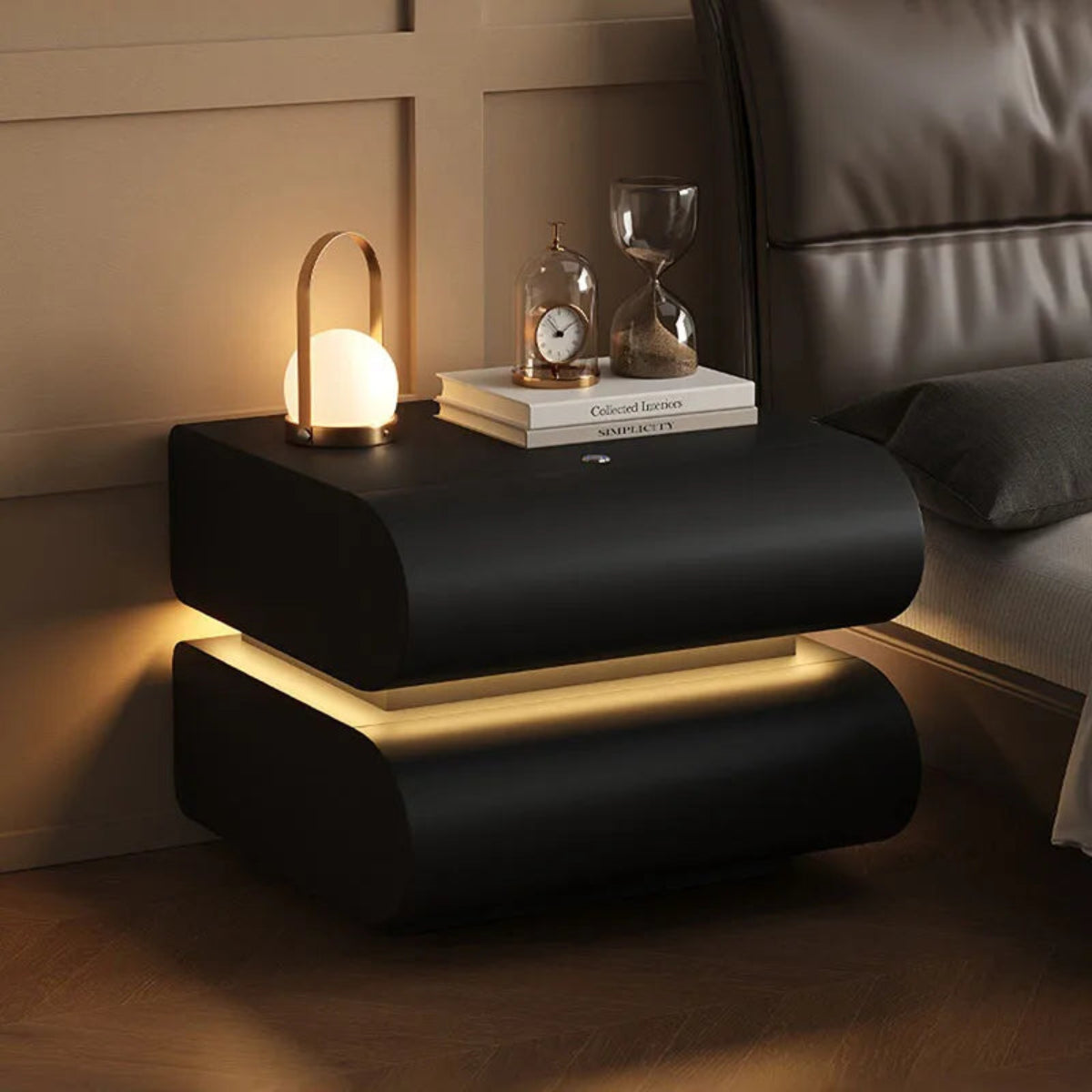 Aura Smart LED luxury nightstand in black with warm LED strip glow styled with a portable lamp, clock, and hourglass beside a dark leather bed in a warm luxury bedroom — LivDecor
