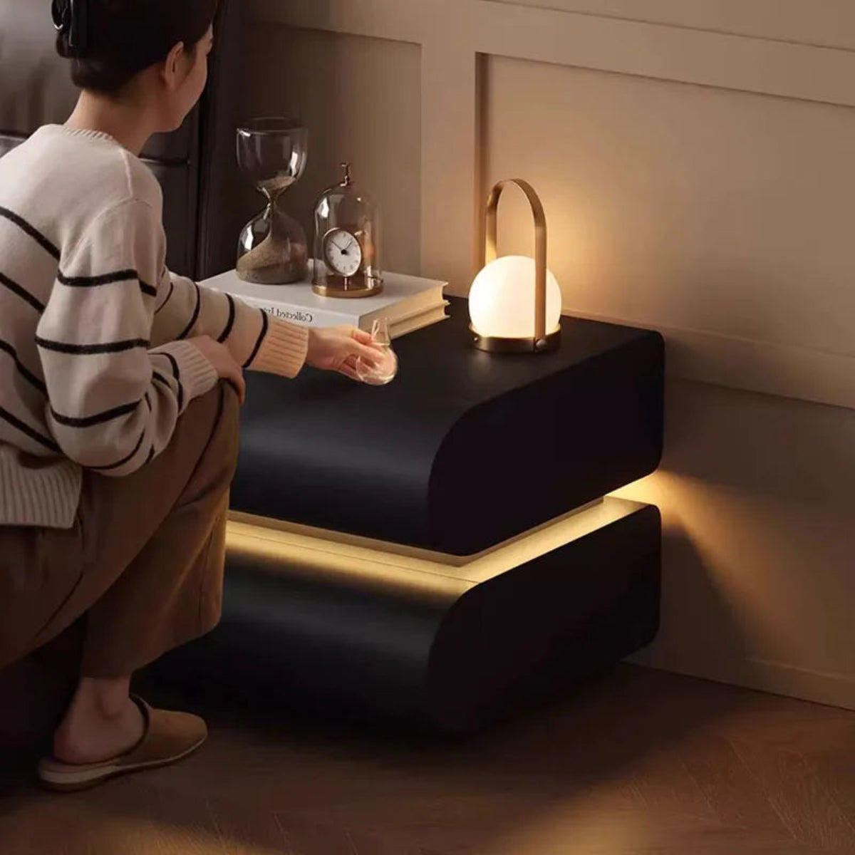 Aura Smart LED luxury nightstand in black with warm LED strip glow and model crouching beside it touching the drawer in a warm luxury bedroom styled with a portable lamp and clock — LivDecor
